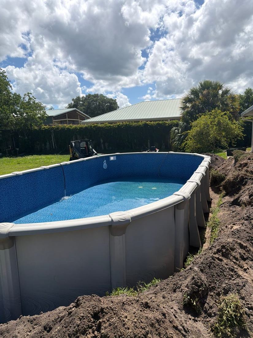 Oval semi-inground pool set down into the cut and filling with water, backfill still open around it